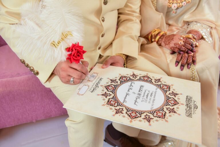 Close-up of a South Asian groom signing the Nikah contract with an ornate feather pen. The bride sits beside him, her hands decorated with henna and adorned with gold jewelry. Both are dressed in traditional light-colored wedding attire, with the contract featuring intricate patterns and Arabic script.