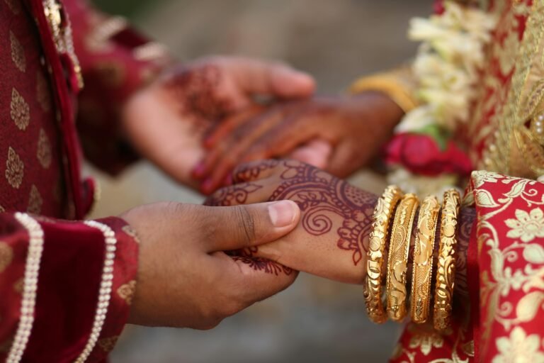 Close-up of a South Asian bride and groom holding hands during a wedding ceremony. The bride's hands are adorned with intricate henna designs and gold bangles, while both wear traditional red and gold embroidered attire.